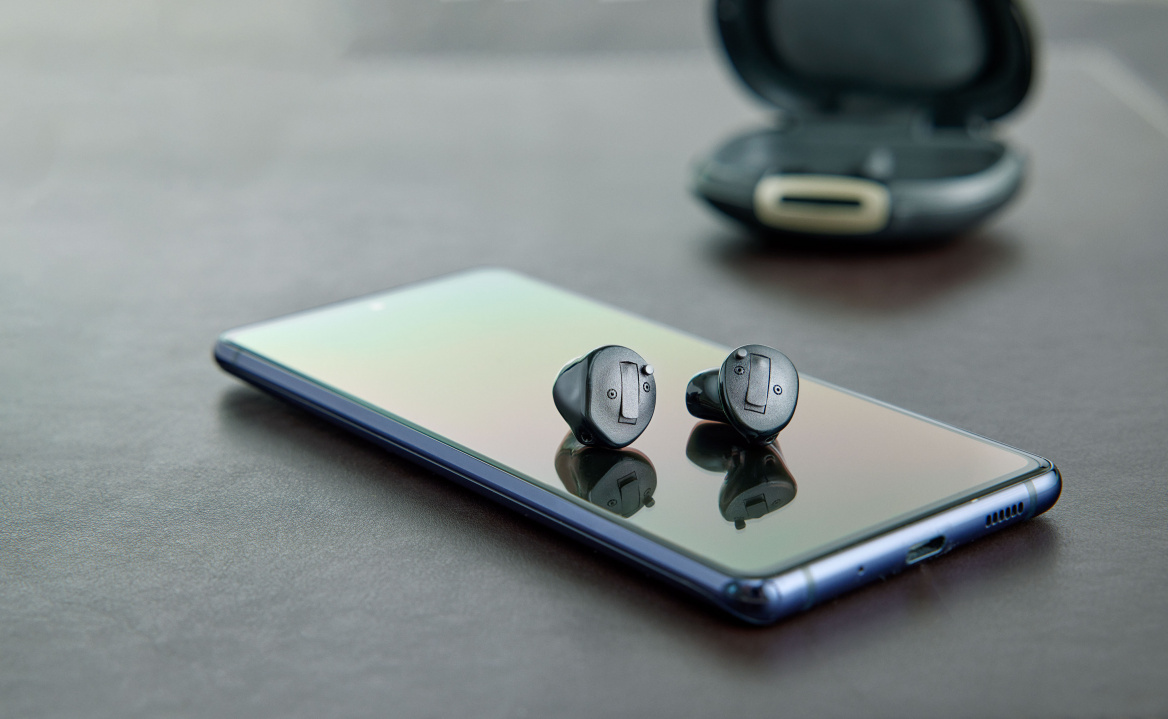 Zwei kleine, schwarze Bluetooth-Hörgeräte liegen auf einem Smartphone, mit einem Ladecase im Hintergrund.
