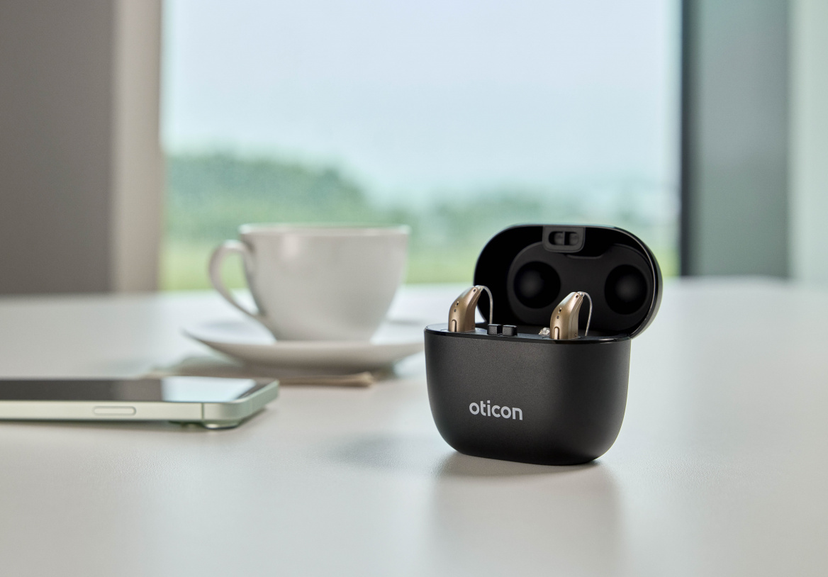 Oticon Intent Hörgeräte in einem schwarzen Ladeetui auf einem Tisch, neben einer weißen Kaffeetasse und einem Smartphone.