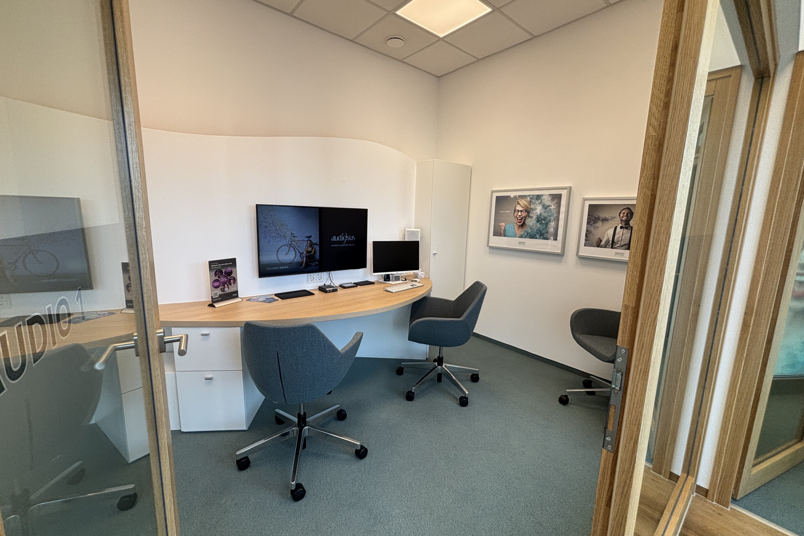 Moderner Beratungsraum bei Hörgeräte Möckel in Saalfeld mit drei ergonomischen Stühlen, einem geschwungenen Schreibtisch, zwei Monitoren und Bildern zufriedener Kund:innen an der Wand.