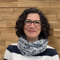 Christine Heubner steht vor einer holzverkleideten Wand und lächelt freundlich in die Kamera. Sie trägt eine schwarz-weiß gestreifte Bluse, eine Brille und einen hellen Schal mit floralem Muster.