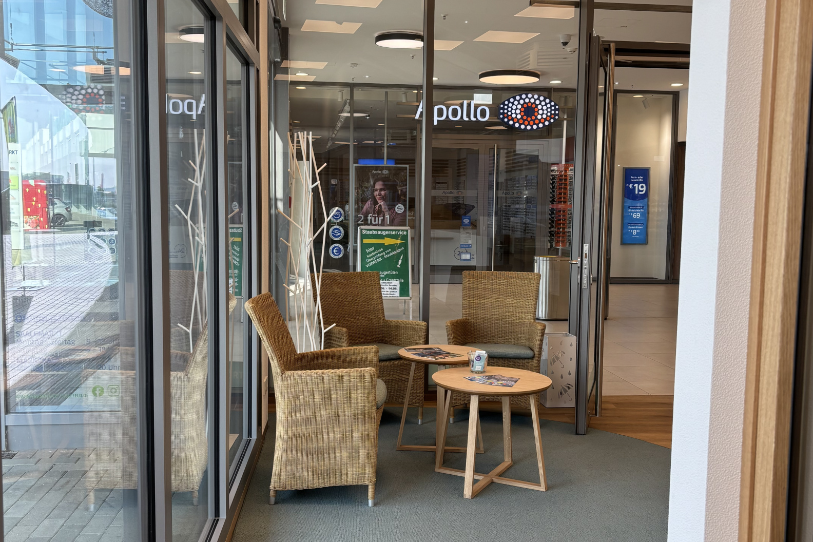 Ein einladender Wartebereich bei Hörgeräte Möckel Saalfeld mit vier Rattanstühlen, zwei runden Holztischen und Blick auf das Nachbargeschäft Apollo hinter Glaswänden.