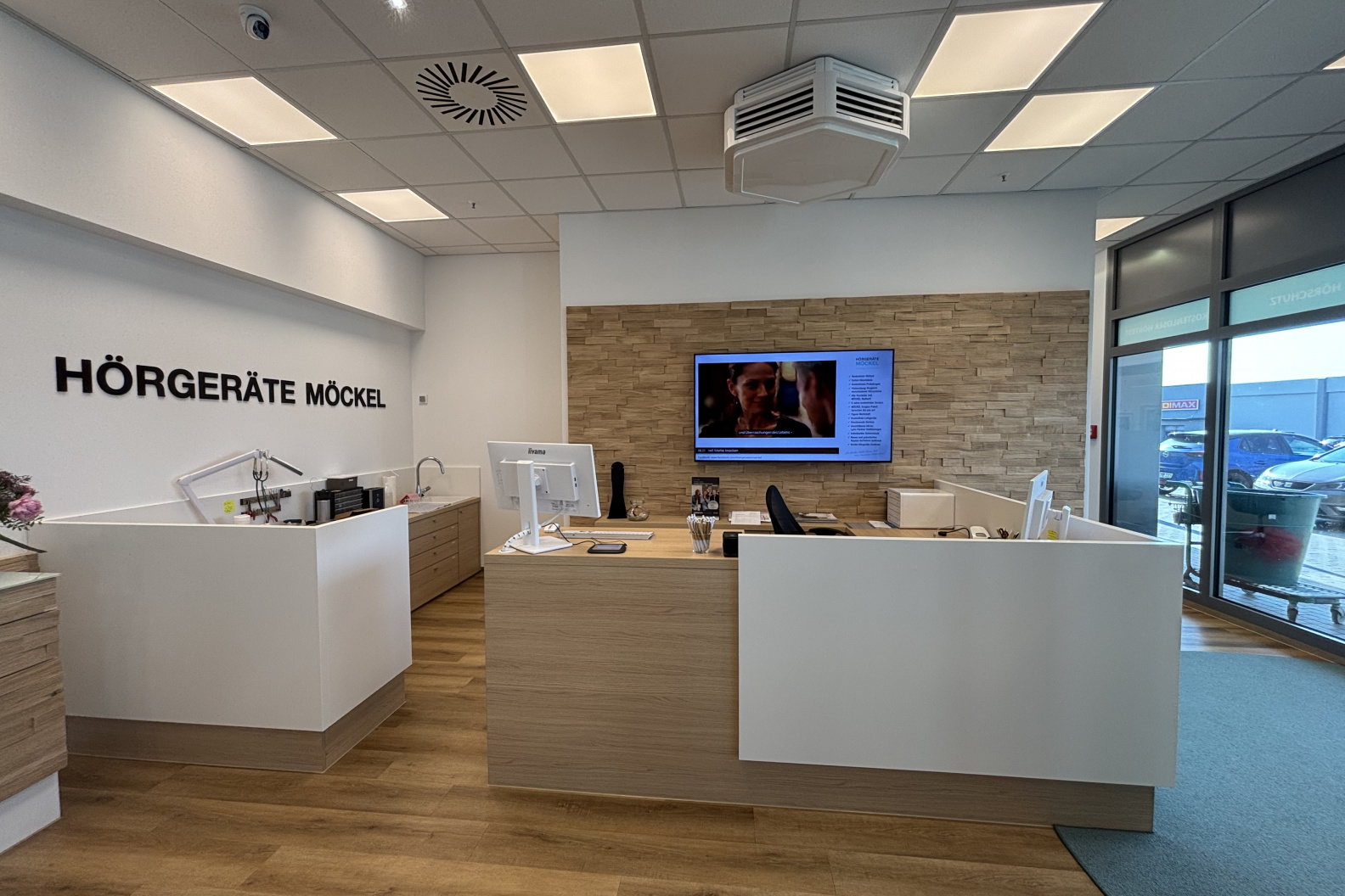 Empfangsbereich von Hörgeräte Möckel in Saalfeld mit zwei weißen Beratungstresen, Holzverkleidung, großem Bildschirm an der Wand und Blick auf den Parkplatz durch die Glasfassade.