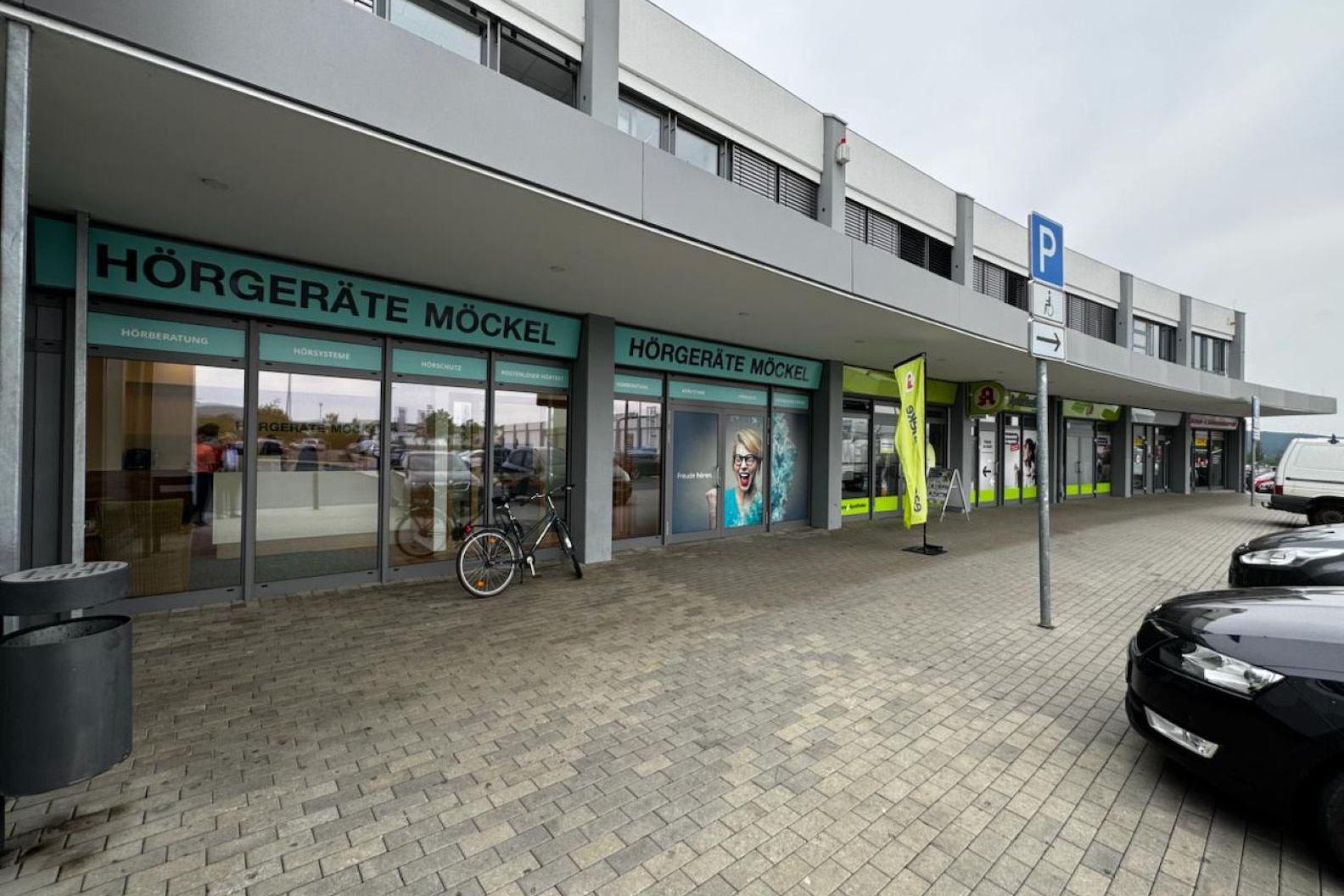 Außenansicht des Hörgeräte Möckel Fachgeschäfts in Saalfeld mit breiter Fensterfront, Fahrrad vor dem Eingang und angrenzenden Geschäften in einem modernen Einkaufszentrum.