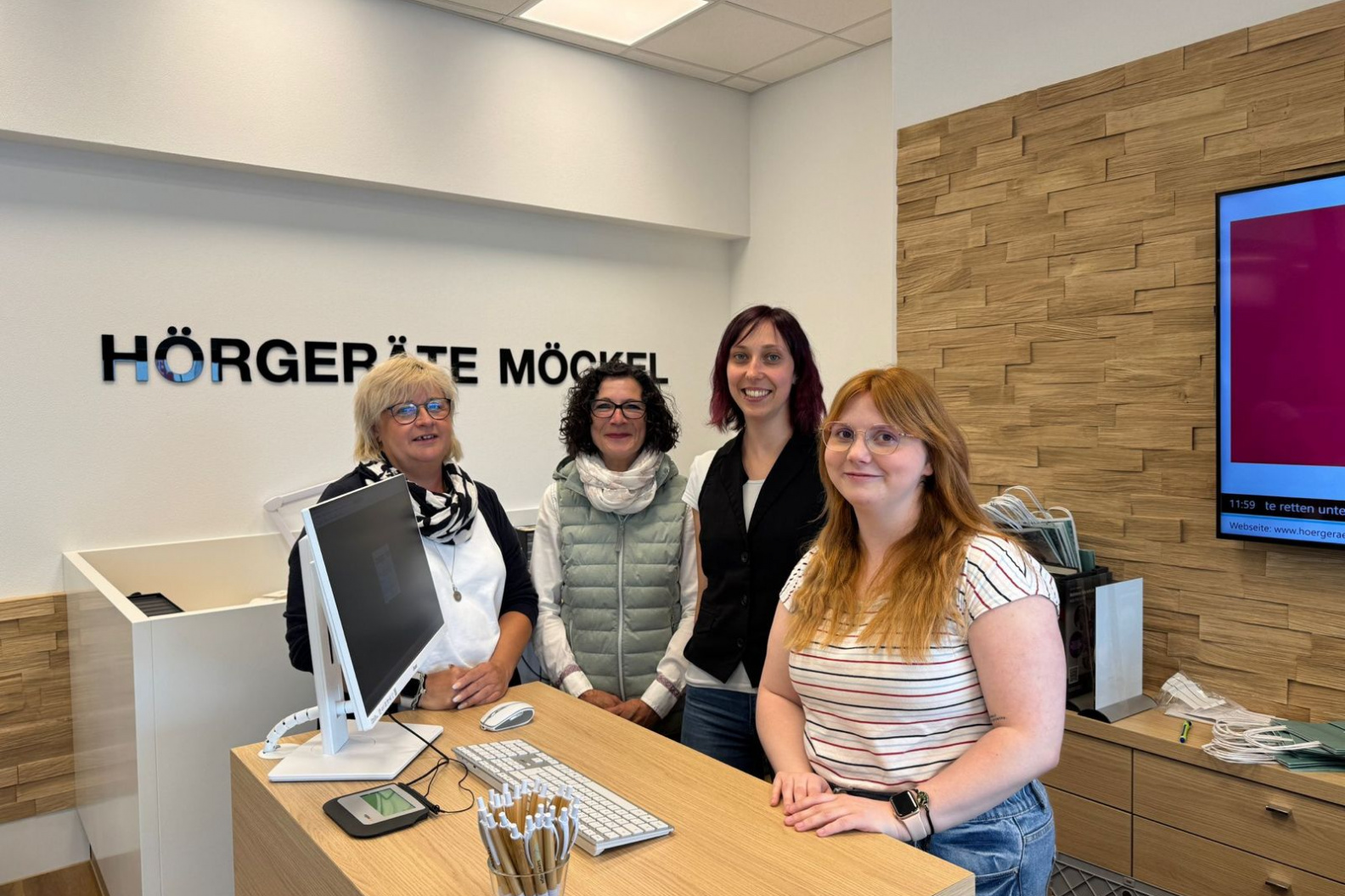 Vier Frauen des Teams von Hörgeräte Möckel Saalfeld stehen lächelnd hinter dem Empfangstresen. Im Hintergrund ist das Firmenlogo „Hörgeräte Möckel“ an der Wand angebracht, rechts ein großer Bildschirm und ein Holzpaneel.