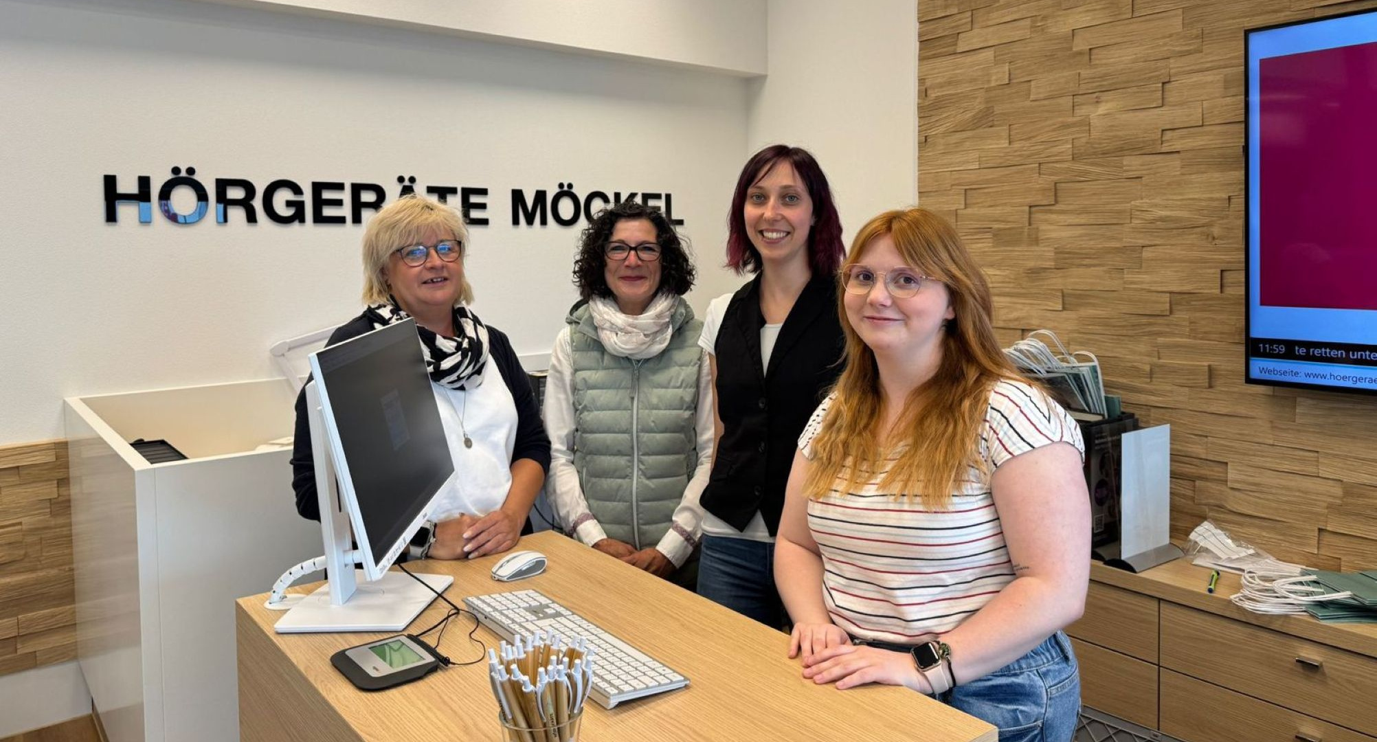 Vier Frauen des Teams von Hörgeräte Möckel Saalfeld stehen lächelnd hinter dem Empfangstresen. Im Hintergrund ist das Firmenlogo „Hörgeräte Möckel“ an der Wand angebracht, rechts ein großer Bildschirm und ein Holzpaneel.