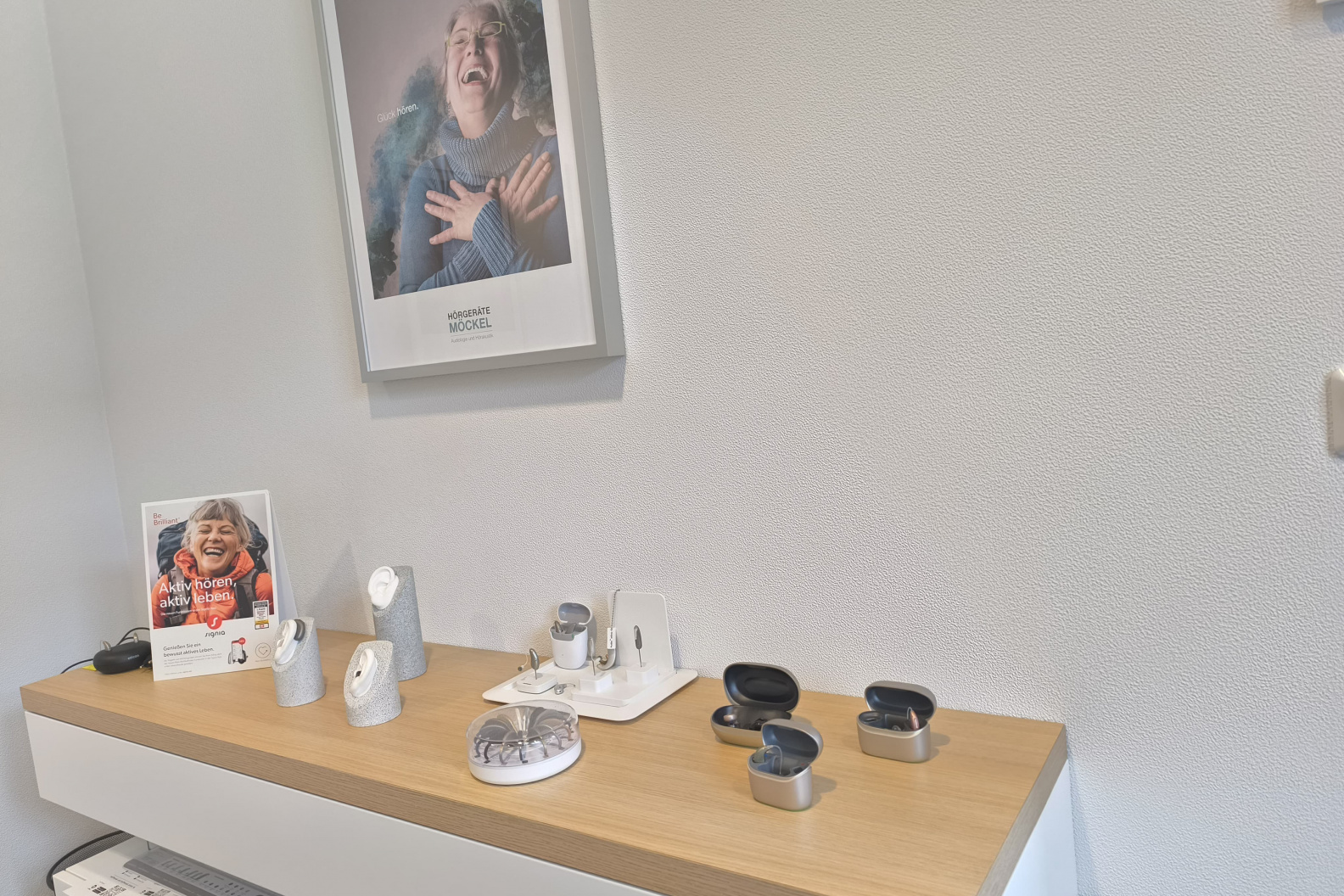 Auslage in der Anpasskabine von Hörgeräte Möckel Weimar mit verschiedenen modernen Hörgeräten und Ladeboxen auf einer Holzablage, im Hintergrund ein Poster mit lachender Frau.