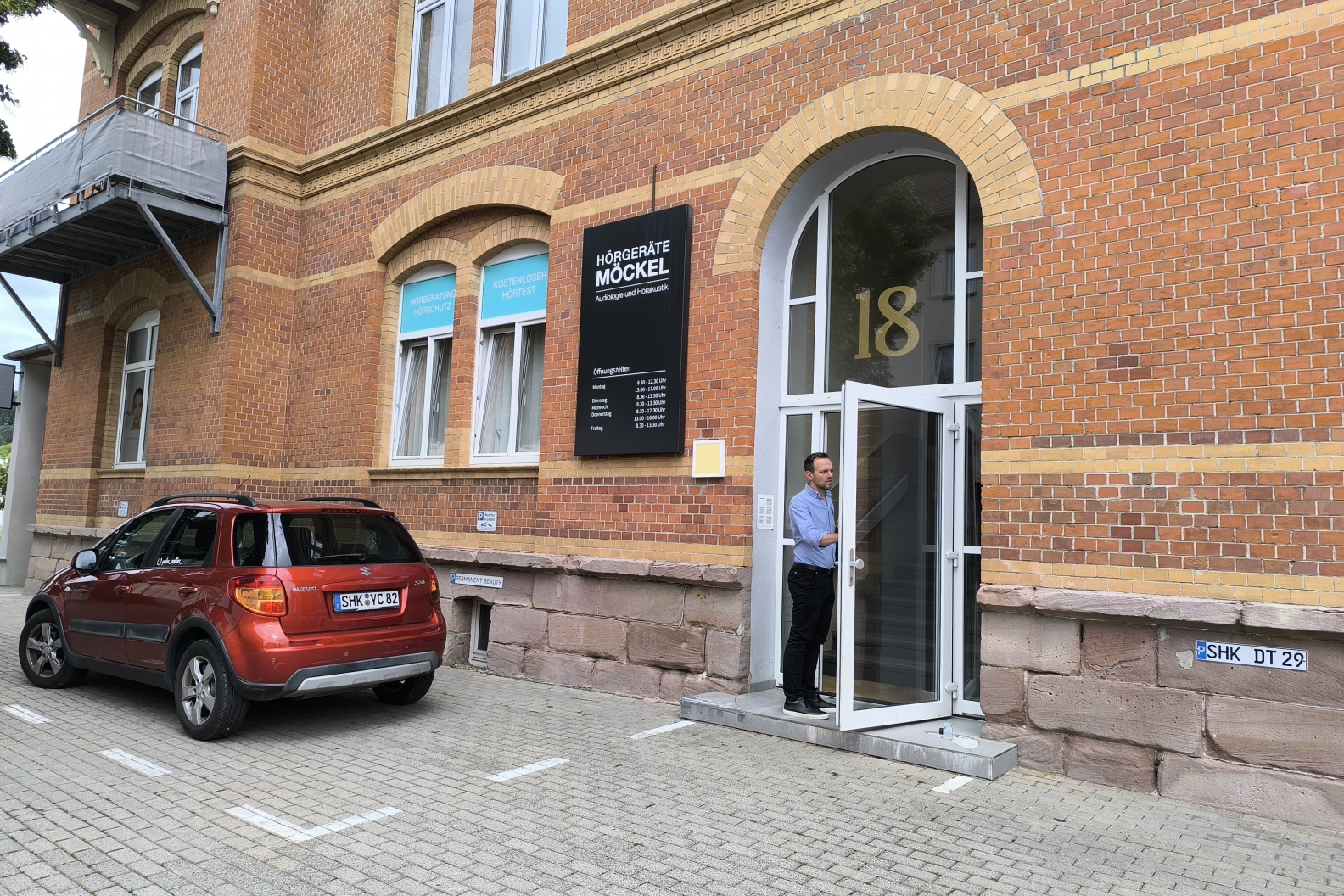 Außenansicht von Hörgeräte Möckel in Kahla, ein Backsteingebäude mit großen Fenstern, schwarzem Firmenschild und geöffneter Glastür mit der Hausnummer 18. Ein Mann steht im Eingang, daneben ist ein rotes Auto geparkt
