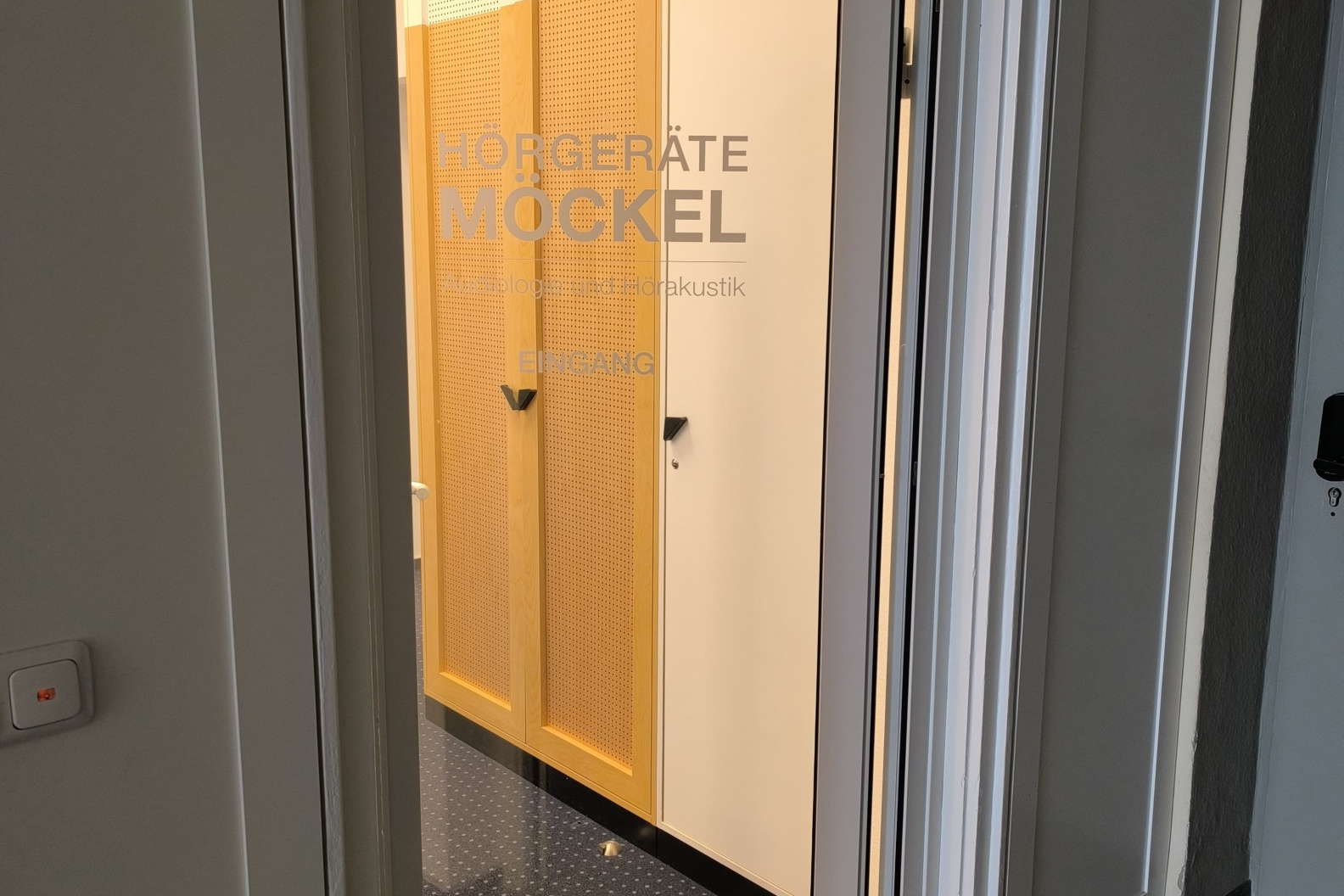 Teilweise geöffnete Glastür mit der Aufschrift „Hörgeräte Möckel“ und „Eingang“, die in einen hellen Raum mit Holzwand und blauem Teppichboden führt.