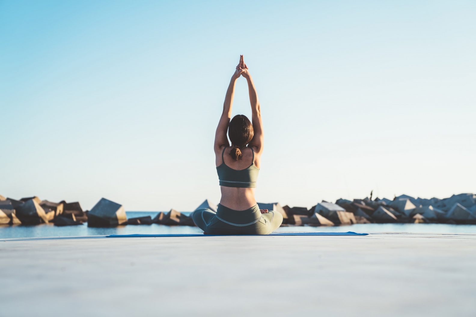 Frau sitzt im Schneidersitz auf einer Yogamatte am Wasser, streckt die Arme nach oben und genießt die ruhige Meereskulisse.