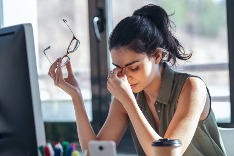 Junge Frau am Schreibtisch massiert ihre Nasenwurzel und hält eine Brille in der Hand, sichtbar gestresst oder von Kopfschmerzen geplagt – Symbolbild für Tinnitus und Hörgeräte mit Tinnitus-Noiser.