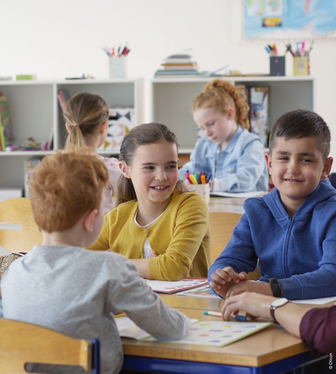 Kinder sitzen gemeinsam im Klassenzimmer am Tisch, lächelnd im Gespräch, Symbolbild für pädakustische Hörtests in der Filiale.
