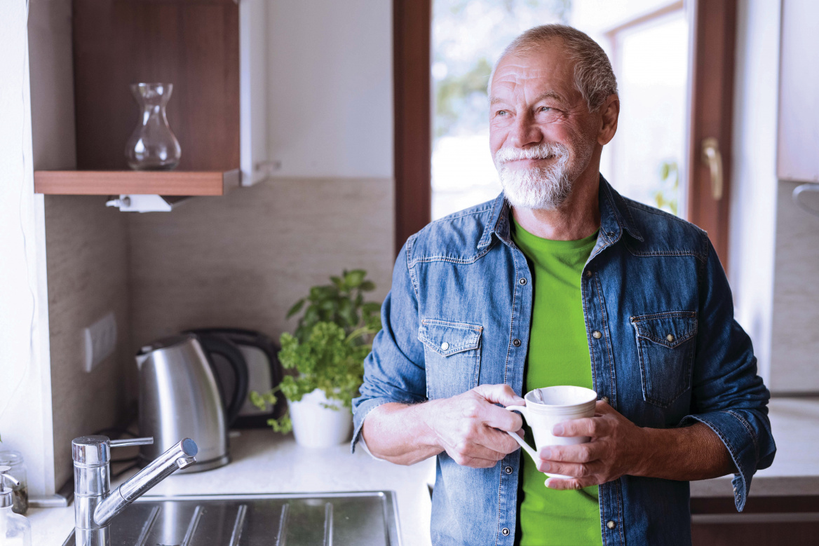 Älterer Mann mit grauem Bart steht lächelnd in einer hellen Küche, hält eine weiße Kaffeetasse und schaut nachdenklich aus dem Fenster – Symbol für Zufriedenheit und Lebensqualität nach einem Hörtest in der Filiale.