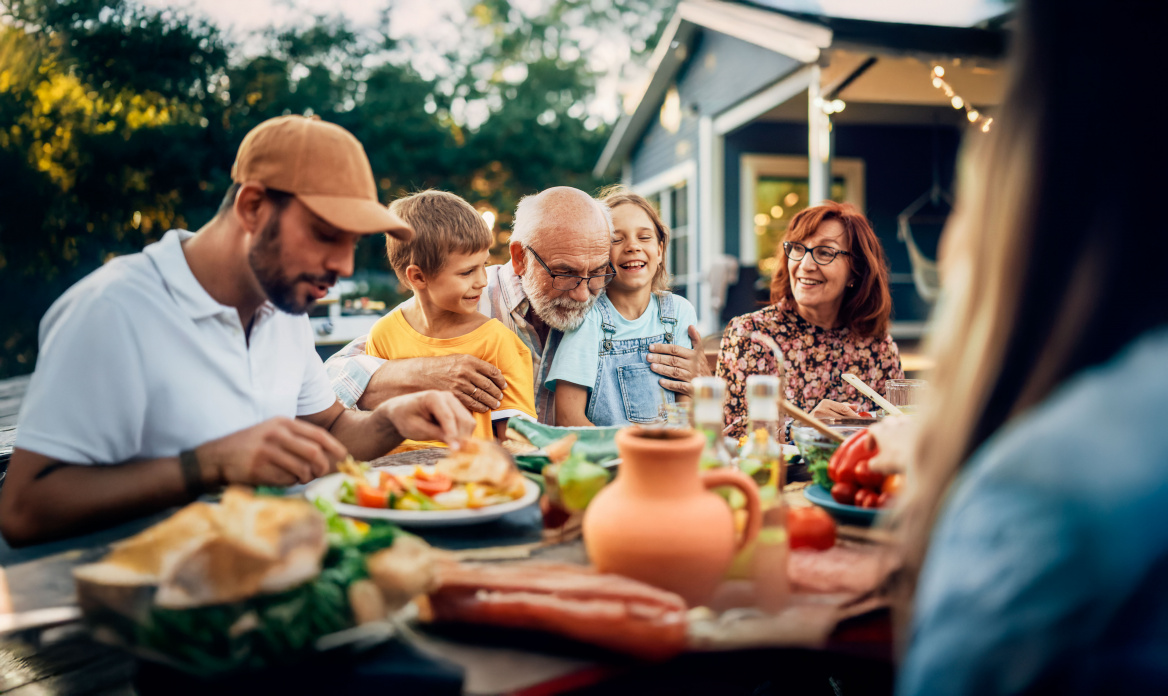 Eine mehrteilige Familie mit Großeltern und Enkelkindern sitzt fröhlich an einem reich gedeckten Tisch im Garten vor einem Ferienhaus.