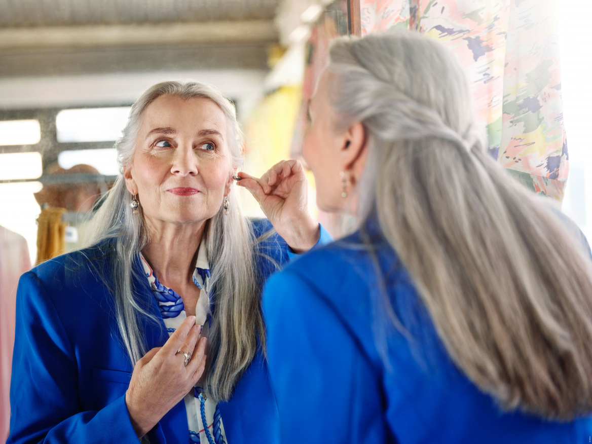 Eine ältere Frau mit langen grauen Haaren und einem blauen Blazer blickt in einen Spiegel, während sie sich mit der Hand ein unauffälliges Im-Ohr-Hörgerät (IIC-Bauform) in ihr Ohr einsetzt.