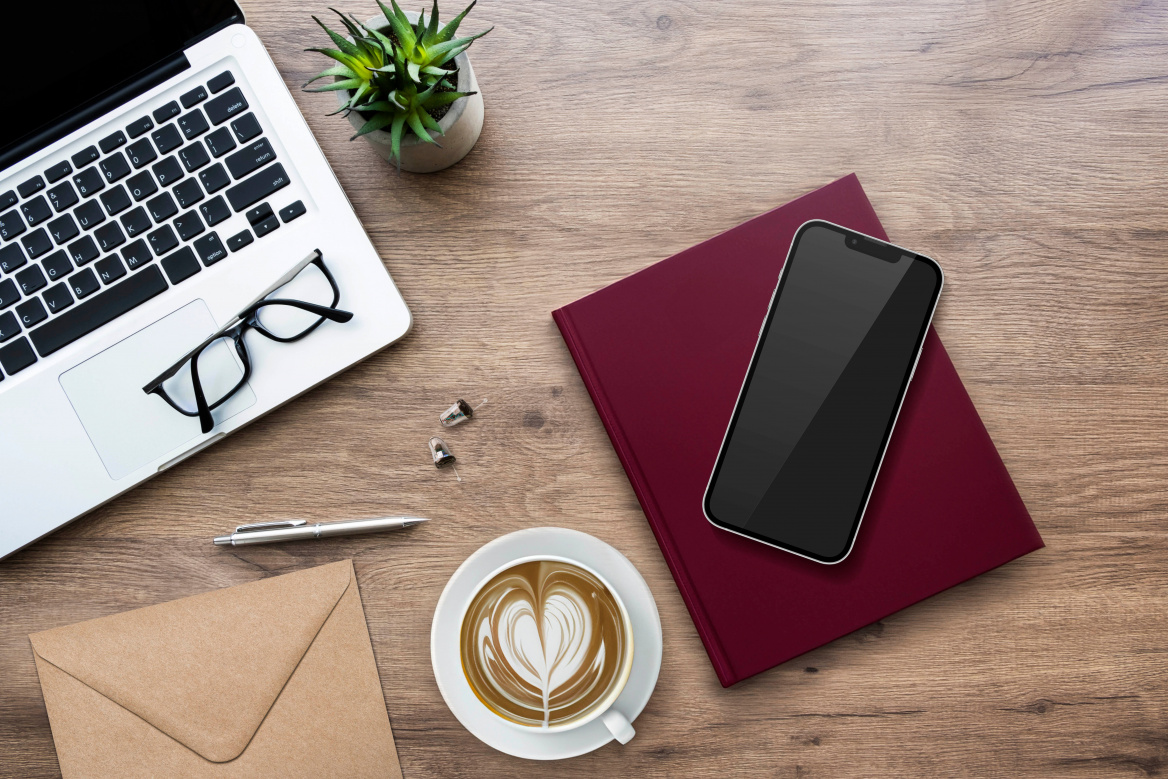 Draufsicht auf einen hölzernen Arbeitsplatz mit einem Laptop, einer Lesebrille, einer Tasse Cappuccino, einem Smartphone auf einem roten Notizbuch und zwei winzigen, unauffälligen Im-Ohr-Hörgeräten auf der Tischplatte.
