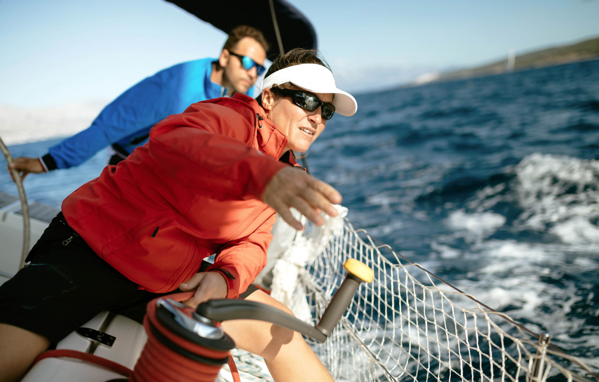 Eine Frau in einer roten Outdoorjacke mit Sonnenbrille und weißer Schirmkappe bedient eine Seilwinde auf einem Segelboot bei windigem Wetter auf dem Meer.