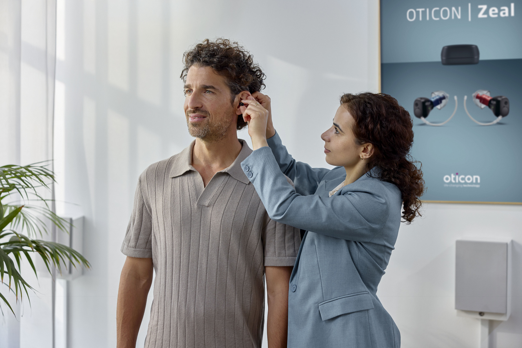 Eine Hörakustikerin setzt einem Mann ein Oticon Zeal Im-Ohr-Hörgerät ins Ohr, während im Hintergrund ein Produktplakat von Oticon Zeal an der Wand hängt.