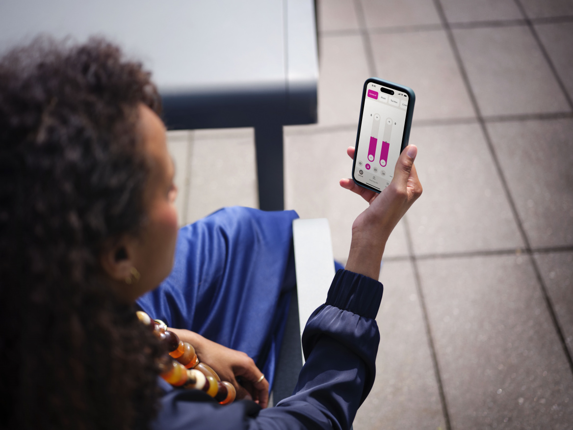 Eine Frau mit dunklen, lockigen Haaren blickt von oben auf ihr Smartphone, auf dem die Oticon Companion App zur Steuerung der Hörgeräte geöffnet ist, während sie auf einer Terrasse sitzt.