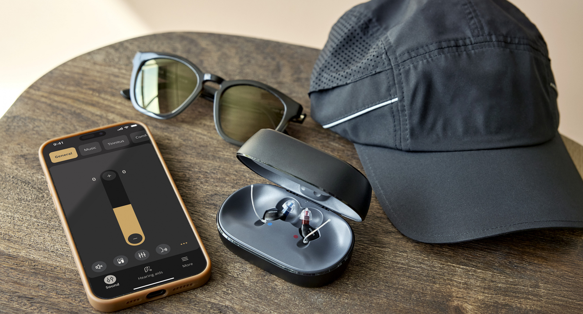 Ein Smartphone mit geöffneter Oticon Companion App, daneben zwei Oticon Zeal Im-Ohr-Hörgeräte in ihrem geöffneten Ladeetui sowie eine Sonnenbrille und eine graue Sportmütze auf einem Holztisch.